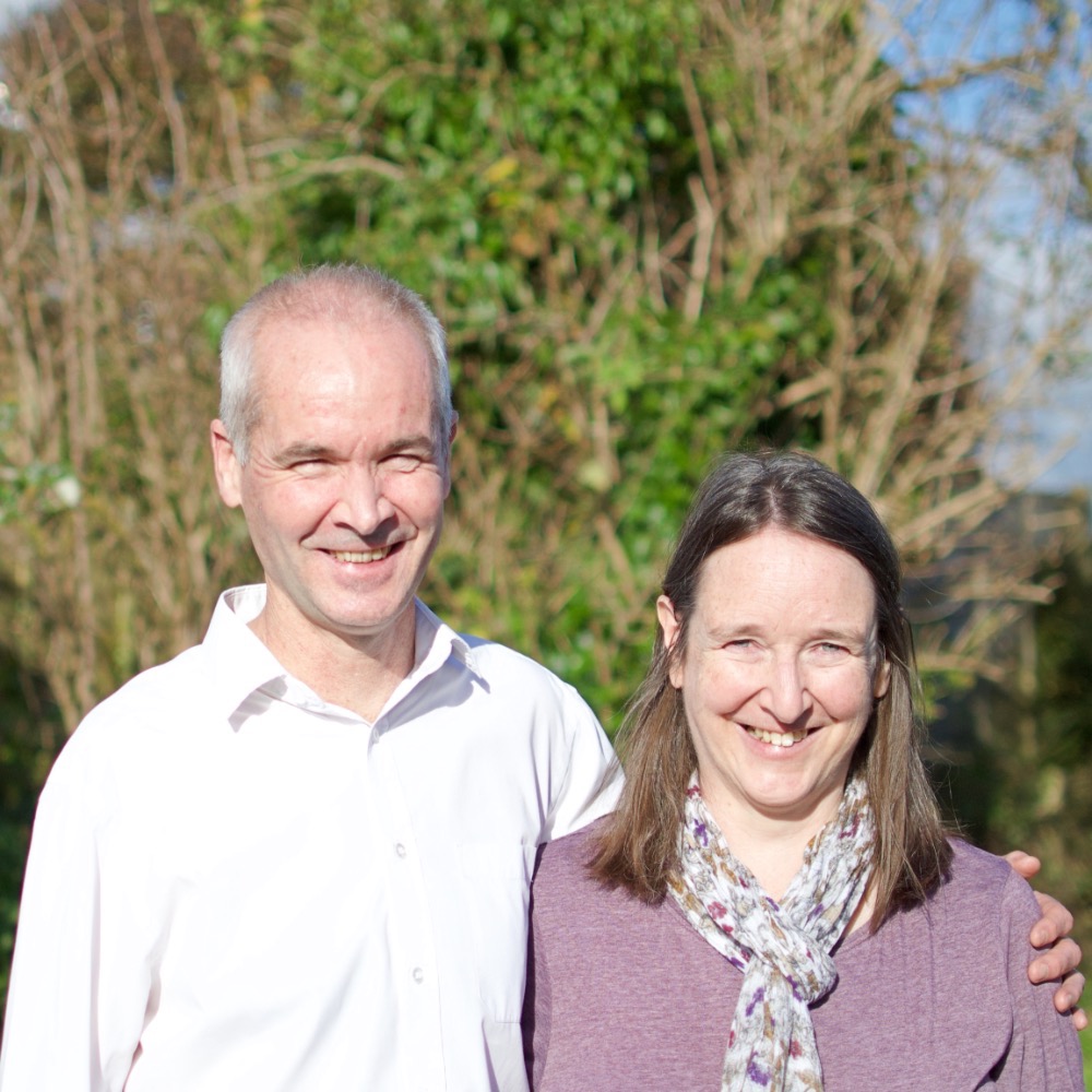 Gavin Walker, minister of Bishopdown Evangelical Church in Salisbury, stands outdoors with his wife Carol. They're smiling against a backdrop of lush greenery and a partly cloudy sky. Gavin is in a white shirt, while Carol complements the scene in her purple top and patterned scarf.