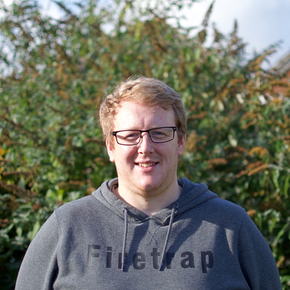 Nick Windust, Assistant Minister at Bishopdown Evangelical In Salisbury in glasses and a grey sweatshirt stands in front of lush green bushes.